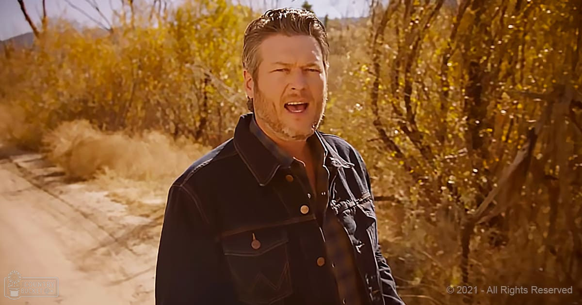 A young adult male stands in a desert-like environment with dry golden bushes, wearing a fitted dark denim jacket over a dark-colored collared shirt or t-shirt. The setting suggests autumnal light and natural daylight.