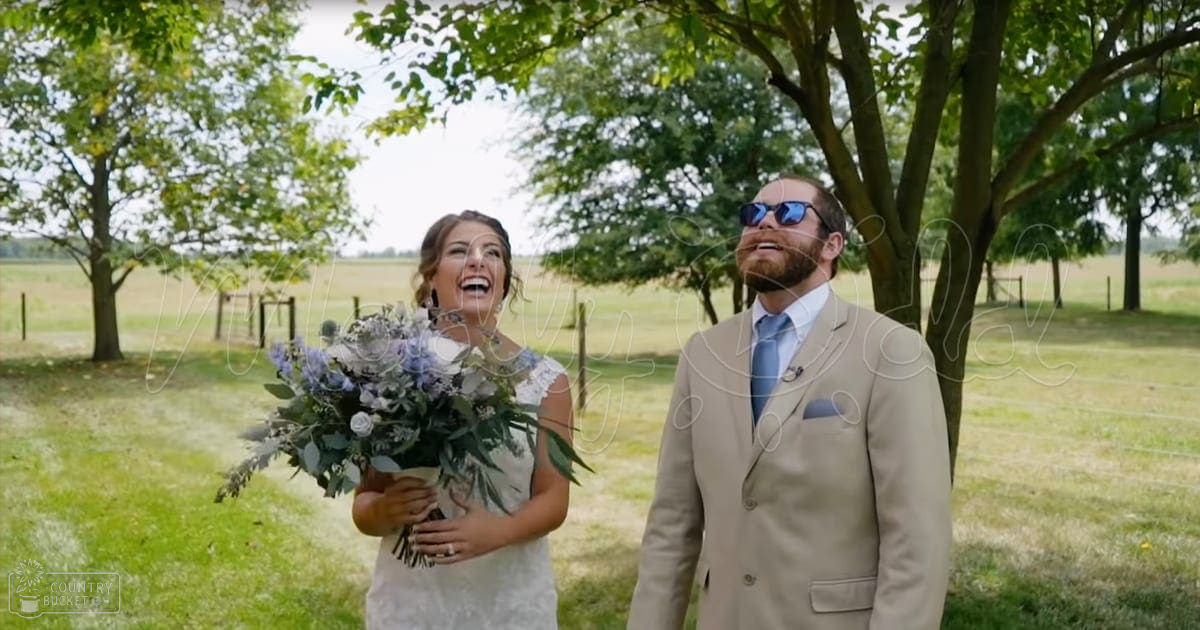 A young adult male and female couple, dressed for a wedding, stand outdoors amidst greenery with trees and a fence, under bright daylight.