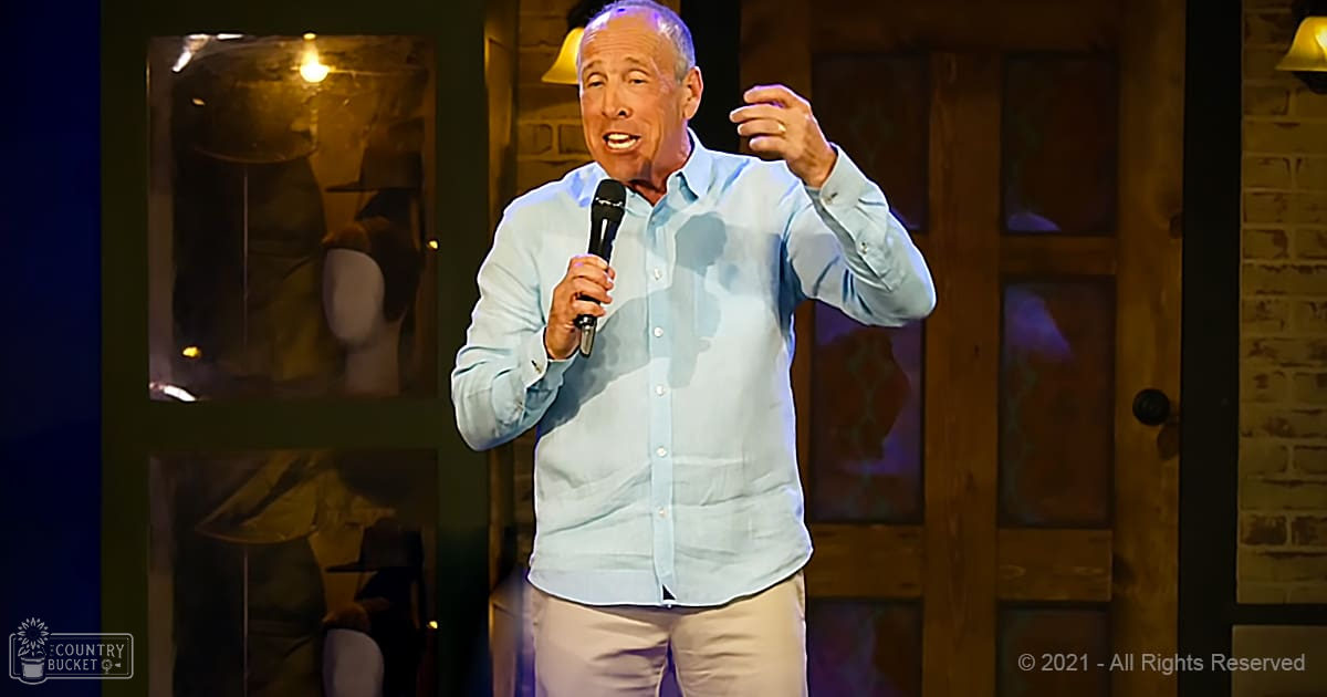 A middle-aged man in a light blue button-down shirt performs stand-up comedy on an indoor stage with wooden doors and wall-mounted lamps.
