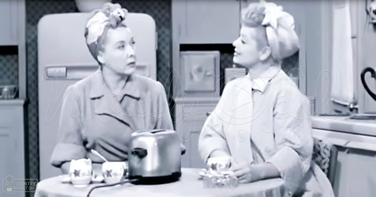 Two women in a retro kitchen setting; one adult female wears a dark blouse with collar and short sleeves, another child-like figure in a light dress with puffed sleeves and high collar, both styled up with large bows or flowers on their hair.