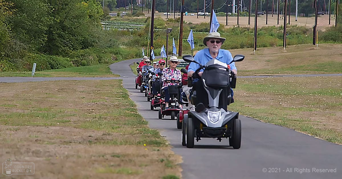 Older adults riding mobility scooters along a grassy pathway with late afternoon light in an outdoor recreational area.