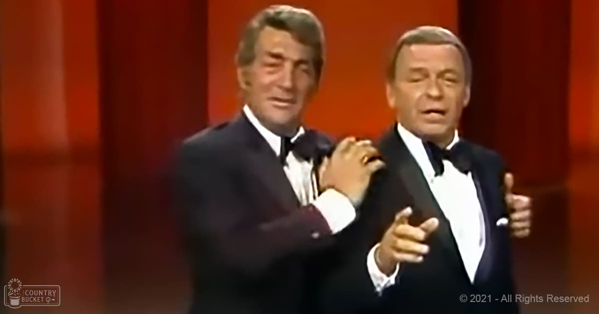 Stage performance with two middle-aged to older adult men dressed formally, possibly from a television show or concert; red curtains and bright stage lighting suggest an indoor venue.