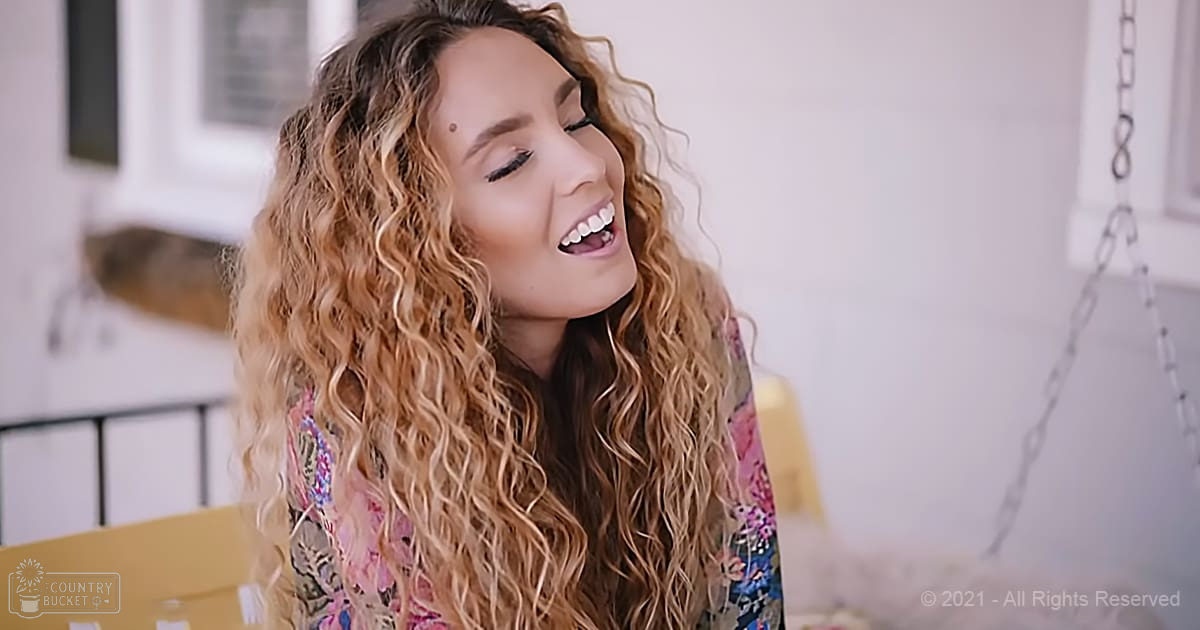 A young adult female wearing a floral-patterned top with long sleeves stands indoors, surrounded by minimal decor including a yellow chair and some indistinct wall art.