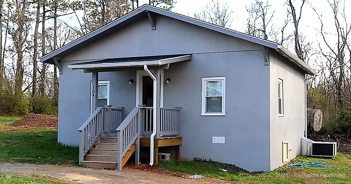 A small house with light gray siding, located outdoors amidst trees, suggesting a rural or suburban environment.