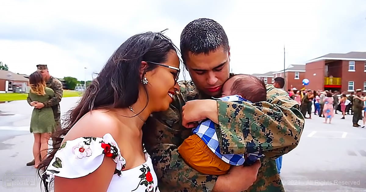 A military couple with their toddler at an outdoor event on a paved area surrounded by grass and buildings under a green and blue sky during spring or summer.