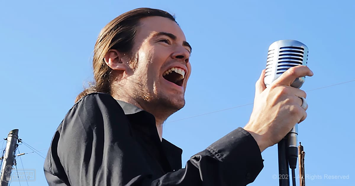 A young adult male sings outdoors under a clear blue sky, wearing a relaxed fit long-sleeved black shirt with a modern casual style.