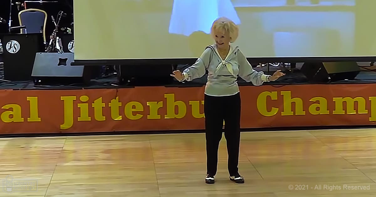 An older adult female in a light gray hoodie and black pants stands on stage at the Jitterbug Championship dance competition under artificial lighting.