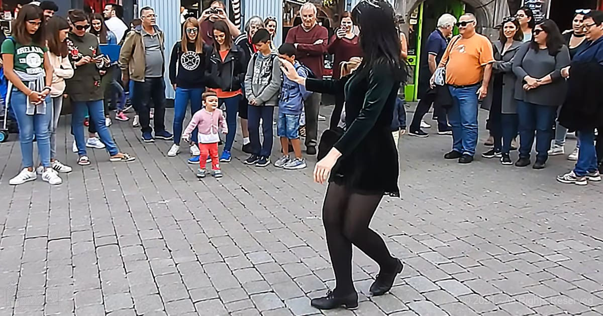 A young adult female dancing energetically on a cobblestone plaza surrounded by an eclectic crowd of onlookers in casual attire.