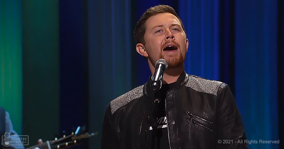 A young adult male singer wearing a black leather jacket with silver accents, performing on an indoor stage with vertical blue curtains and holding a microphone.