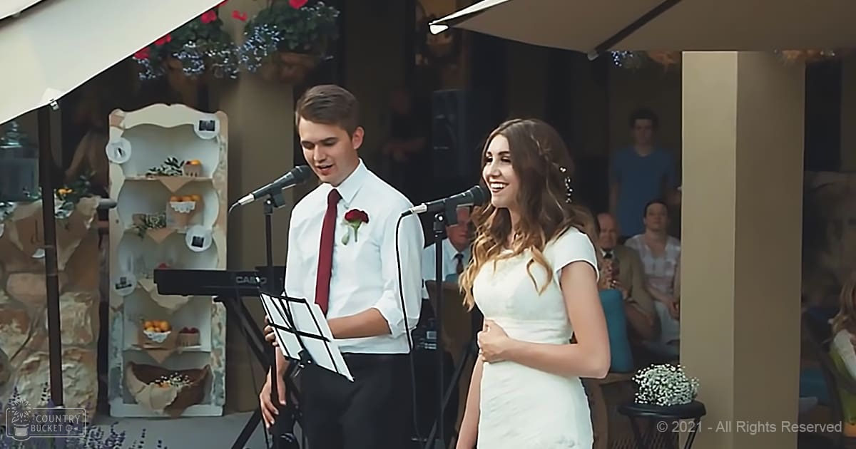 A couple at a wedding ceremony, with the bride wearing a white gown and groom in a white shirt with red tie, seated outdoors under tents or canopies on what appears to be a spring or summer day.