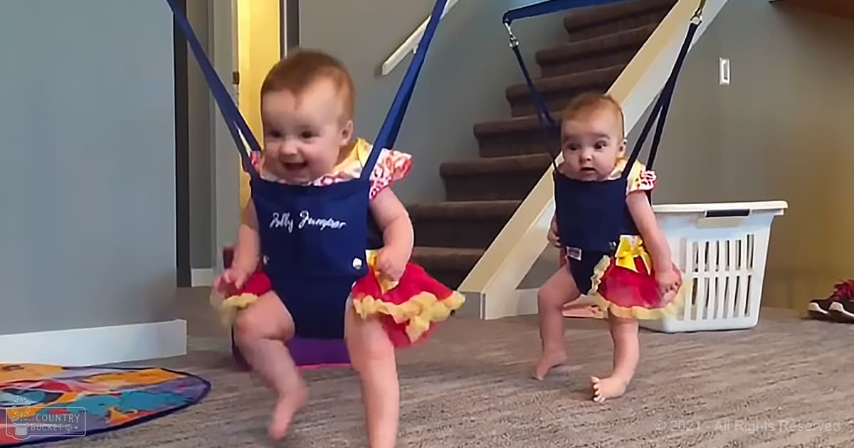 Two infants wearing blue 'Belly Bouncer' garments with ruffled edges and red skirts, standing on a staircase in a family home setting under natural daylight.