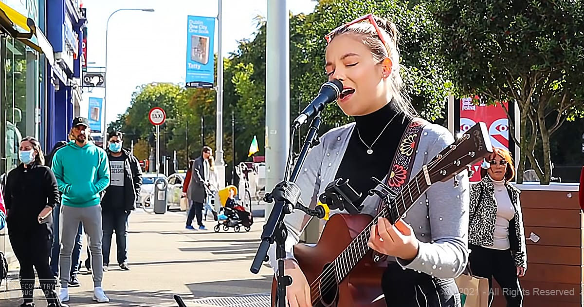 A young adult woman playing a brown acoustic guitar with floral patterns, singing outdoors in an urban setting. She wears a gray cardigan over a black turtleneck and red-framed sunglasses.
