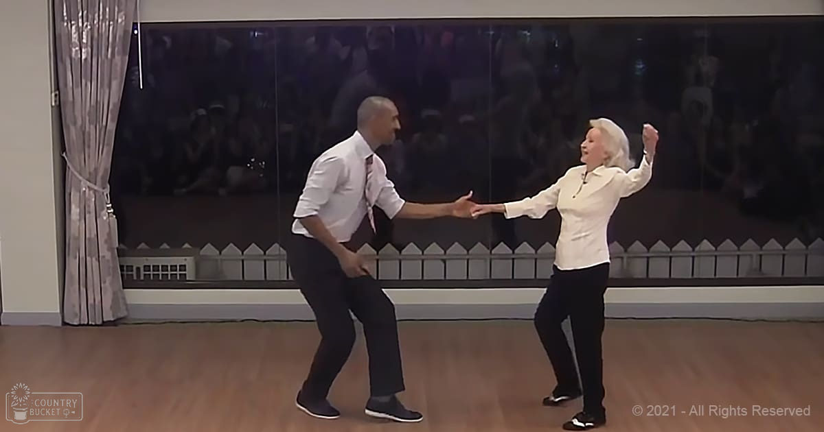 A dance scene in an indoor studio; adult male and older female dancers wearing white tops and dark pants, with a large reflective mirror showing audience reflections.