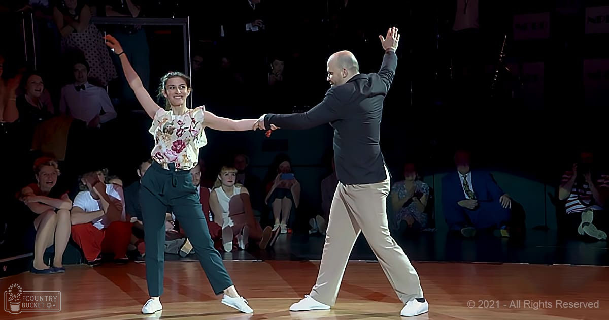 A dance competition scene with a female dancer wearing a floral blouse and pants, performing alongside a male dancer dressed in a blazer and beige pants on an indoor stage under focused lighting.