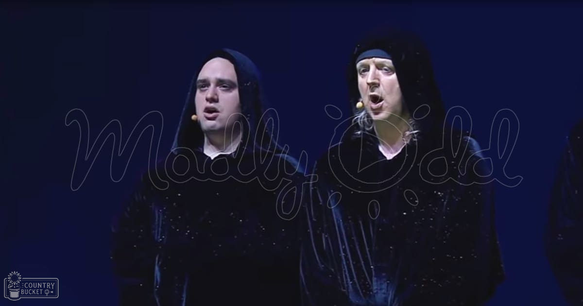 A performance scene with two adult male performers on stage, both wearing black hooded garments with small bead embellishments. The backdrop is dark, suggesting a focused lighting setup typical of theatrical or musical productions.