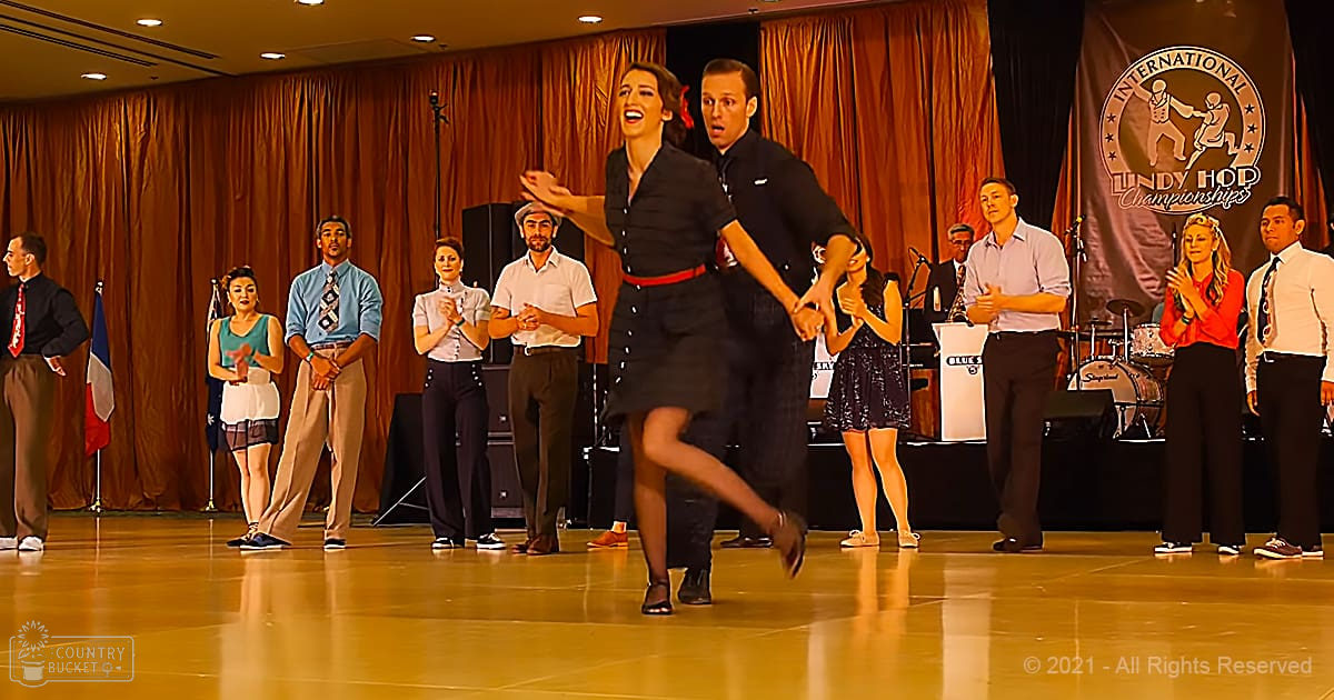 A couple dancing swing in front of an audience at the International Lindy Hop Championships 2021, with a banner reading 'International Lindy Hop Championships,' curtains, and microphones visible.