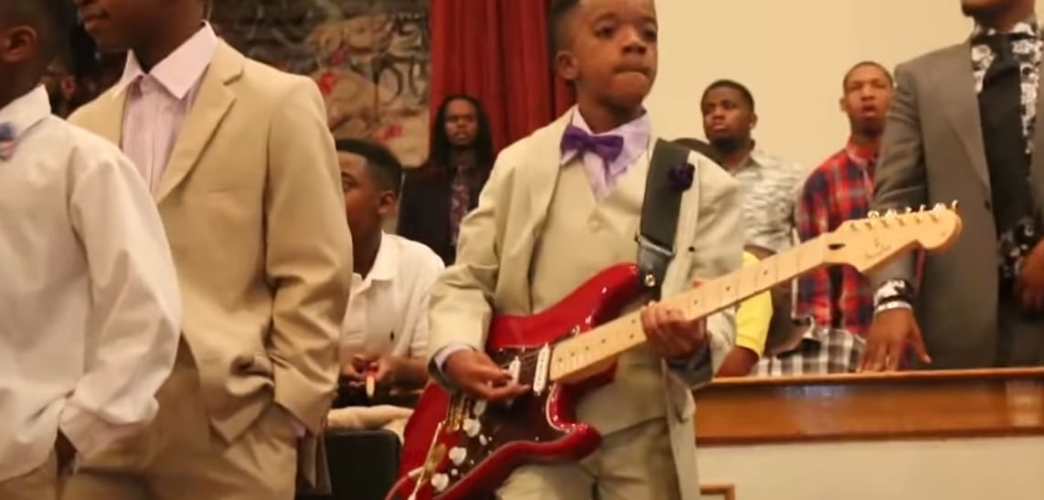 A young boy plays an electric guitar in a church setting with adults and wooden pews partially visible.