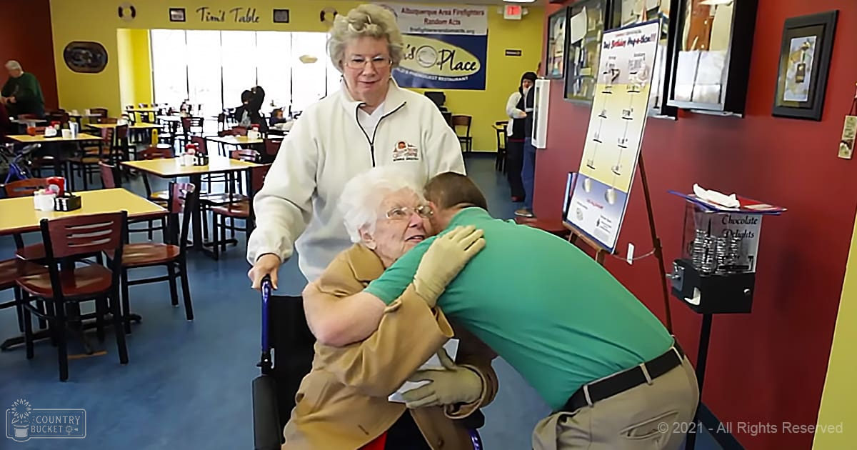 An older adult female interacts with a young adult male in a restaurant setting, surrounded by other patrons and tables with yellow tops.