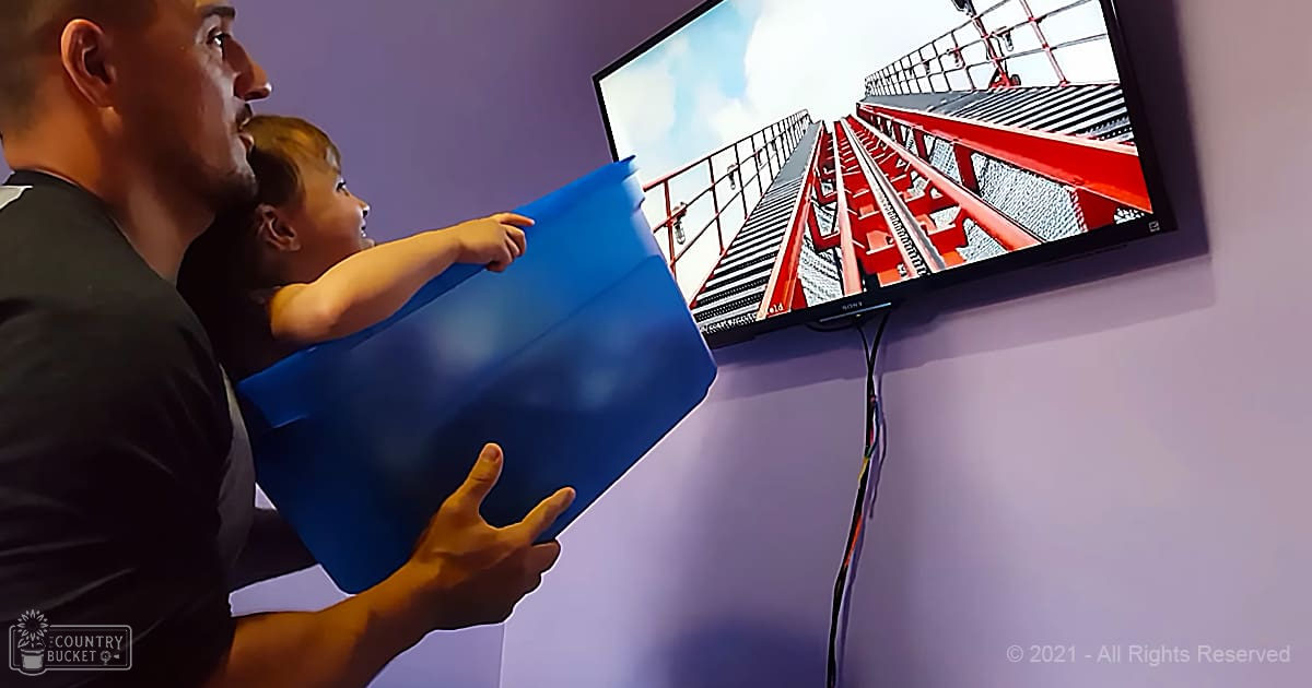 An adult male holding an infant indoors with a wall-mounted television displaying a staircase, blue bucket in the foreground against a purple wall.