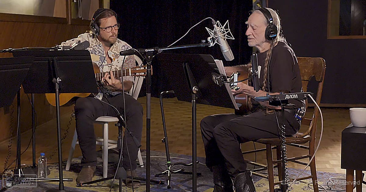 Indoor recording studio setup with two adults engaged in music production; dark background, wooden floor, various musical instruments and equipment present.