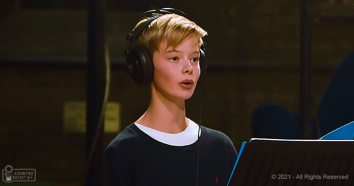 A young adult female wearing a dark blue or black long-sleeved top and large over-ear headphones stands in a recording studio with soundproofing panels on the walls.