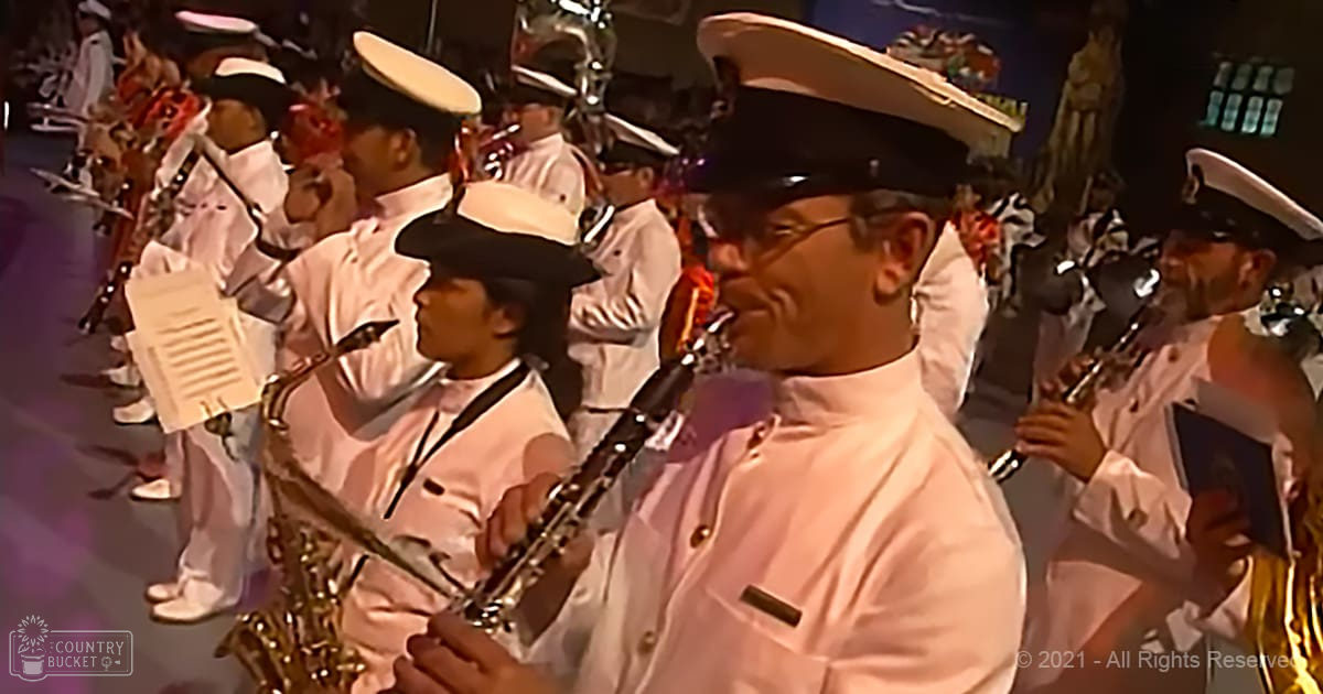 A marching band performance with adult male musicians in white uniforms with black hats, playing clarinets and saxophones indoors under stage lighting.