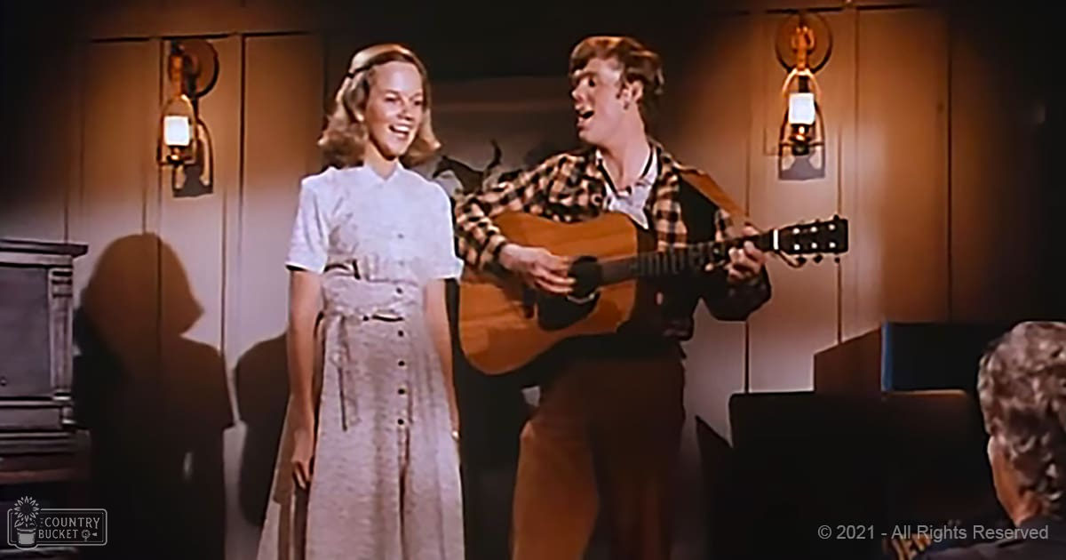 Young adult female wearing a light-colored, mid-20th-century dress with buttons down the front; young adult male in a loose-fitting plaid shirt, playing guitar; indoor setting with wall sconces and piano visible.