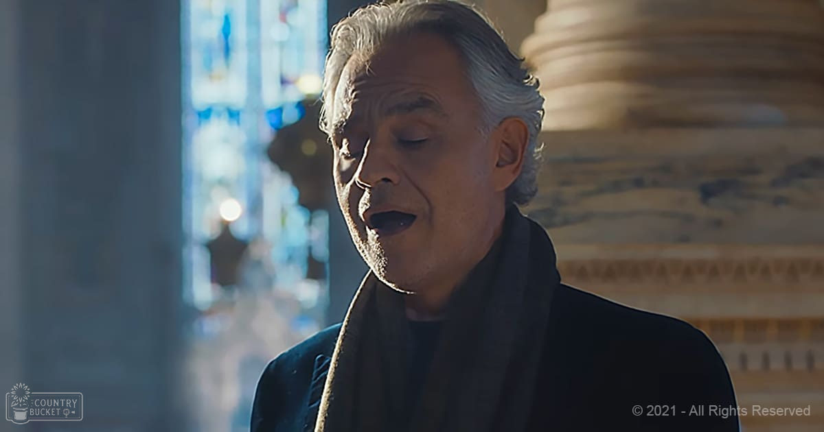 An older adult male singing in a grand interior setting with soft, diffused natural light streaming through large stained glass windows.