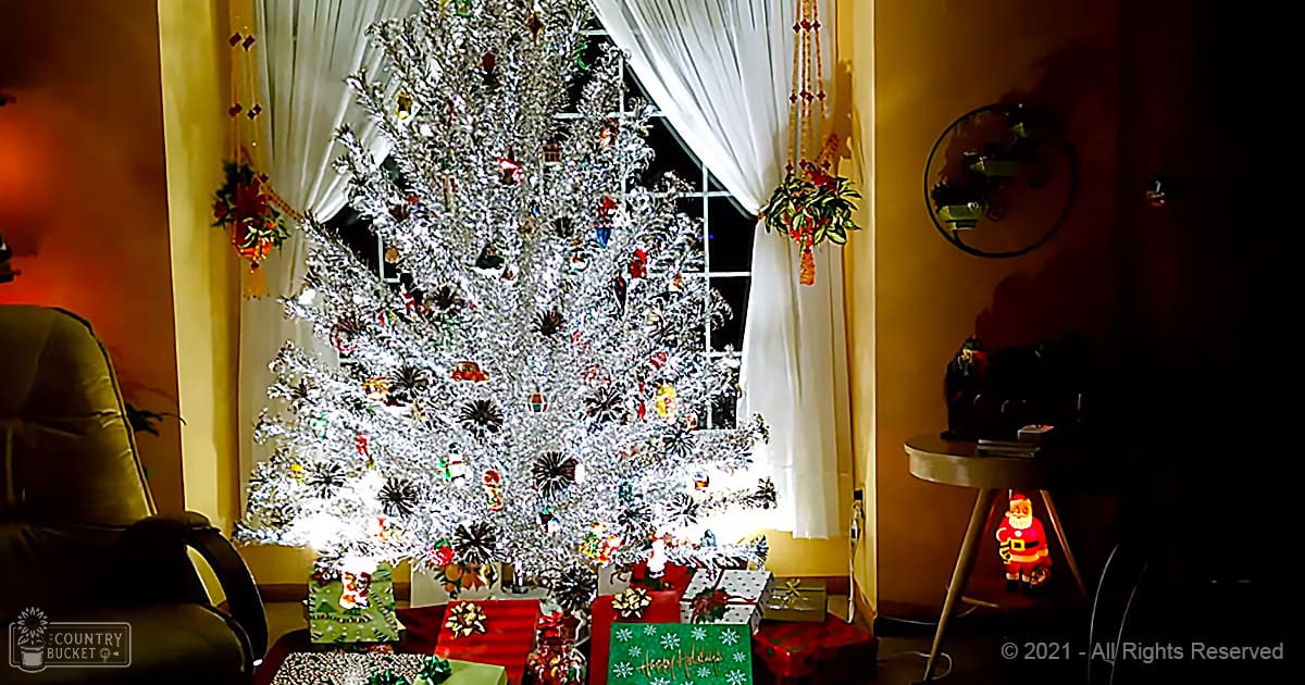 A decorated Christmas tree with ornaments, wrapped presents under it, white curtains, a small round table with a Santa Claus figurine, and a wall clock in an indoor setting.