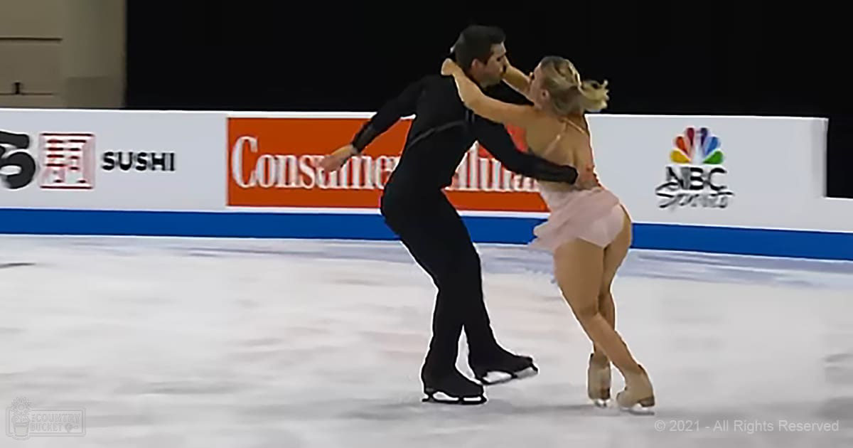 A male adult skater wearing a black long-sleeved outfit and female partner in a light pink dress perform on an indoor ice rink under artificial lighting.