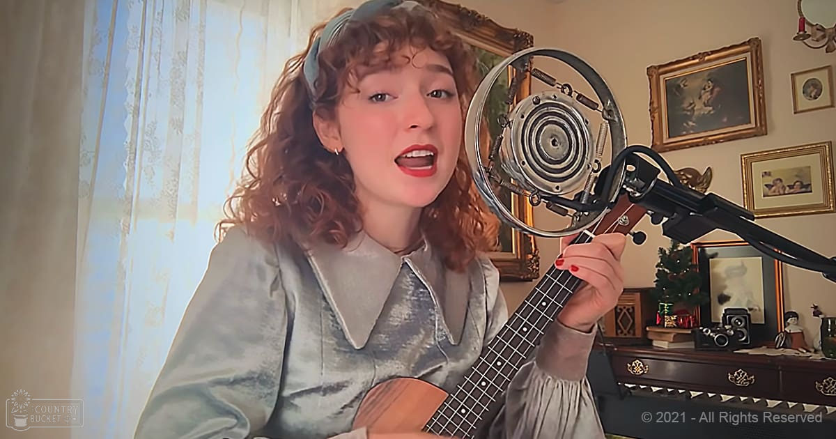 Young adult female singing into a microphone while playing an acoustic guitar in a cozy home setting with natural light, wearing a light blue long-sleeved dress.