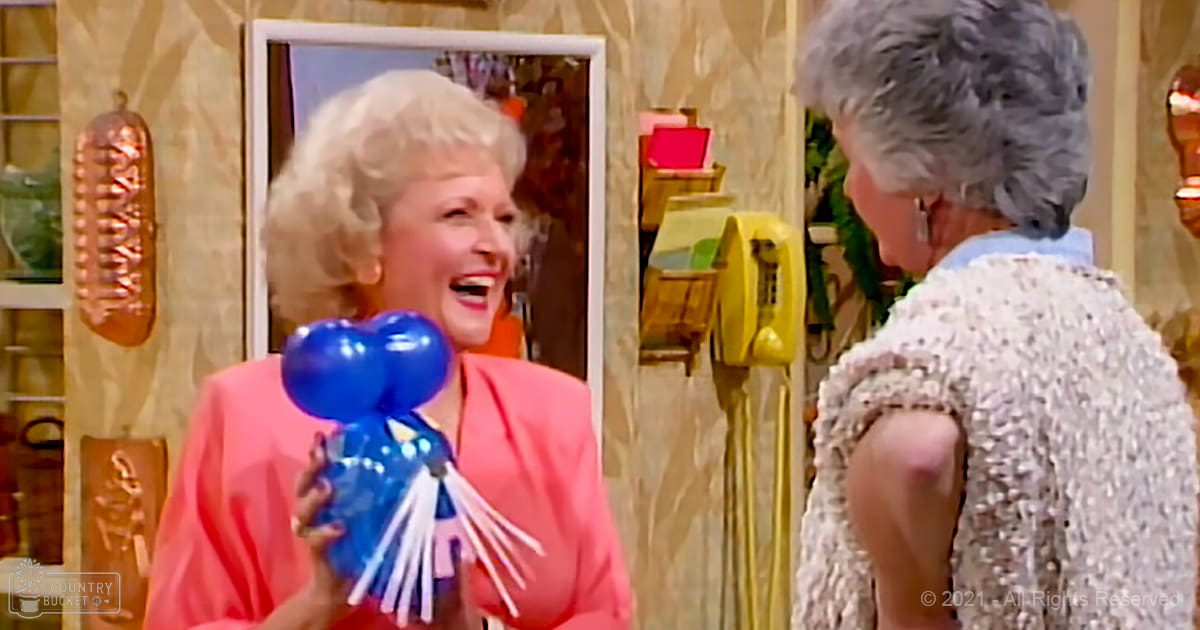 Middle-aged adult female wearing a pink blazer and holding blue balloons with white streamers, accompanied by an older adult female in a white sequined dress, both indoors with vintage decor.