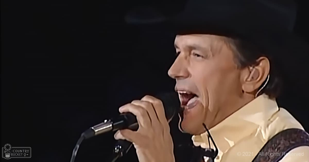 A middle-aged male performer singing into a microphone at an indoor concert setting, wearing a light-colored garment with lace-like details on shoulders in what appears to be stage lighting.