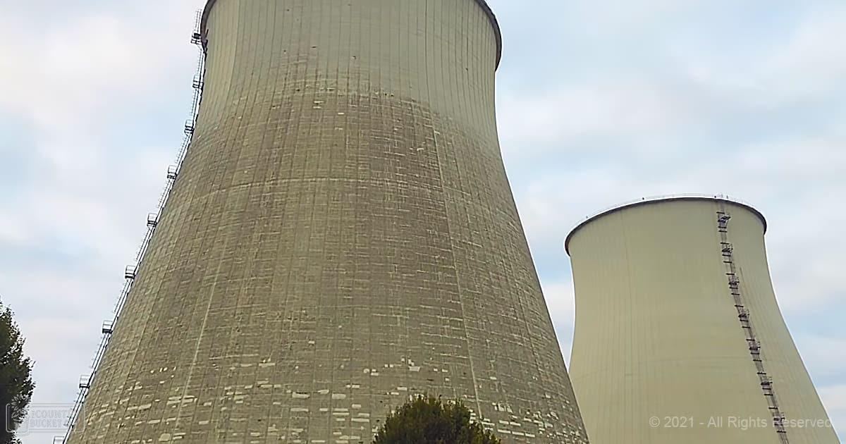 Two large cooling towers with ladders on their sides at a power plant; partly cloudy sky, greenery foreground, daytime light