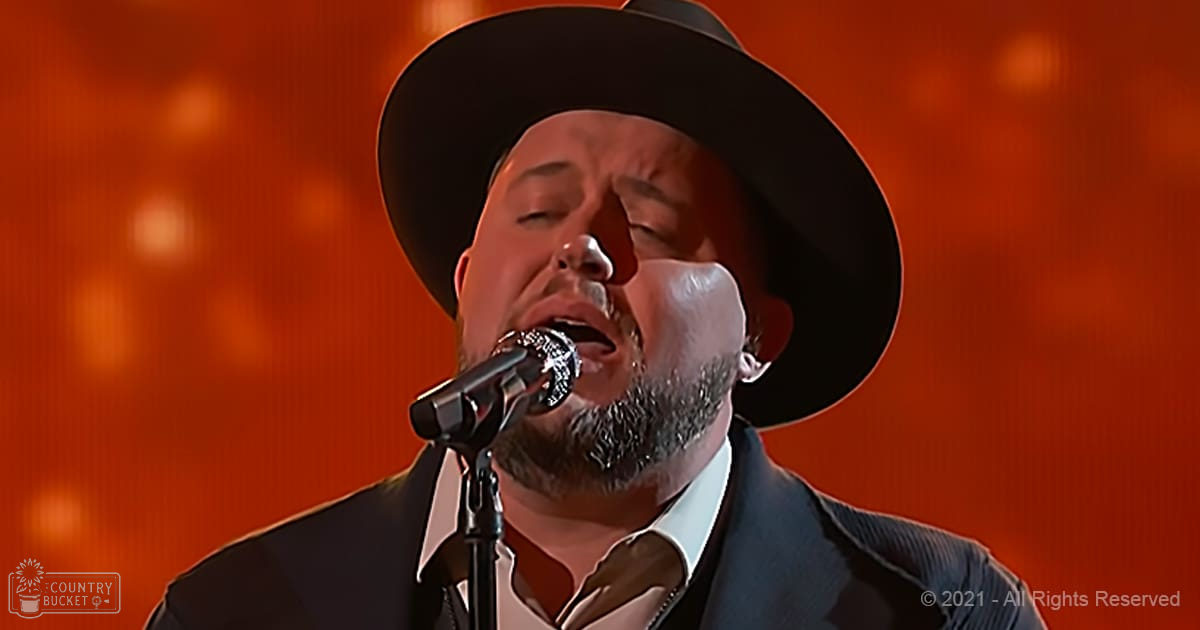 A middle-aged male performer wearing a wide-brimmed black hat, dark suit jacket over an undone white shirt with tie, singing into a microphone on stage under orange lighting.