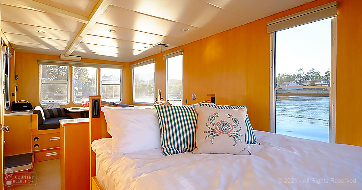 Cozy cabin interior aboard a houseboat with white bedding, striped pillows, and wooden furniture overlooking water and greenery.