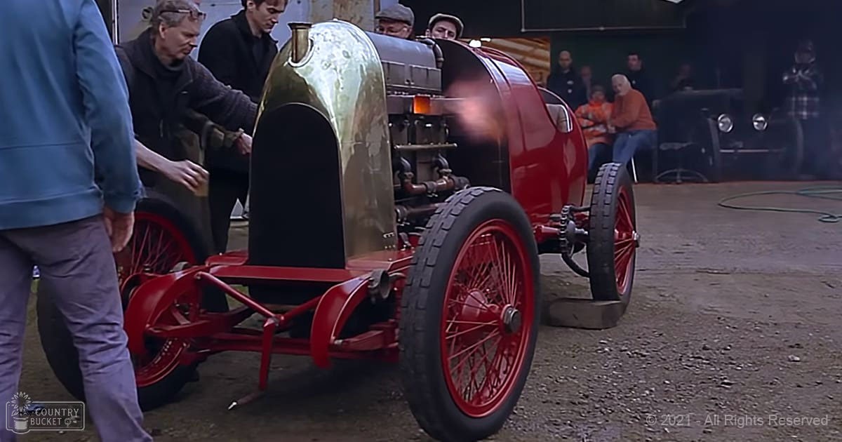 An adult male works on a vintage race car in an indoor workshop with various tools and equipment scattered around. The scene is well-lit, suggesting daytime.