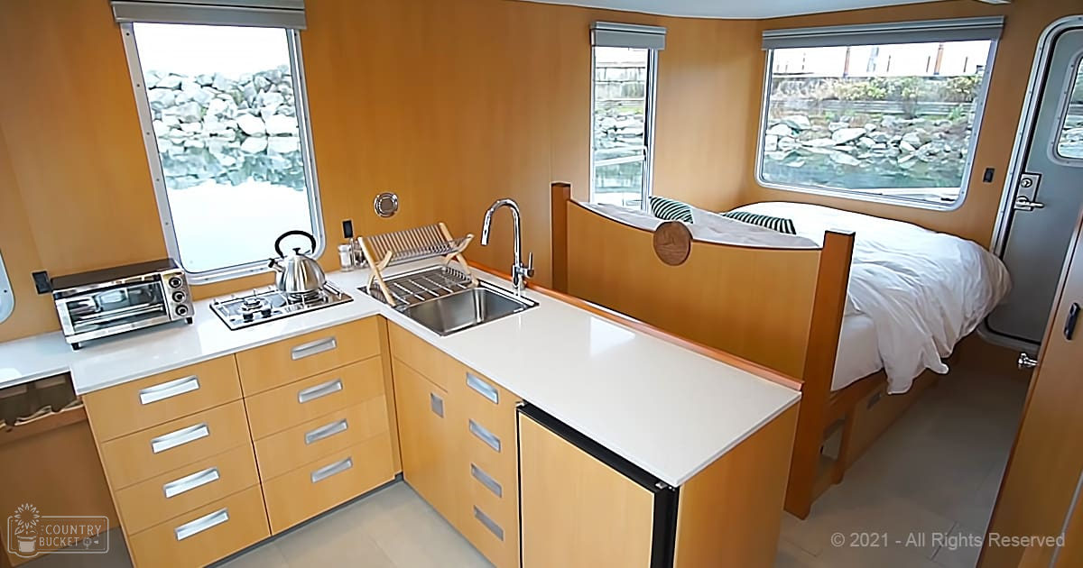 Compact kitchen area in a small RV or mobile home with light wood cabinets, white countertops, stainless steel sink, kettle on stovetop, and bed with wooden headboard. Outdoor view of rocks through windows suggests daytime.
