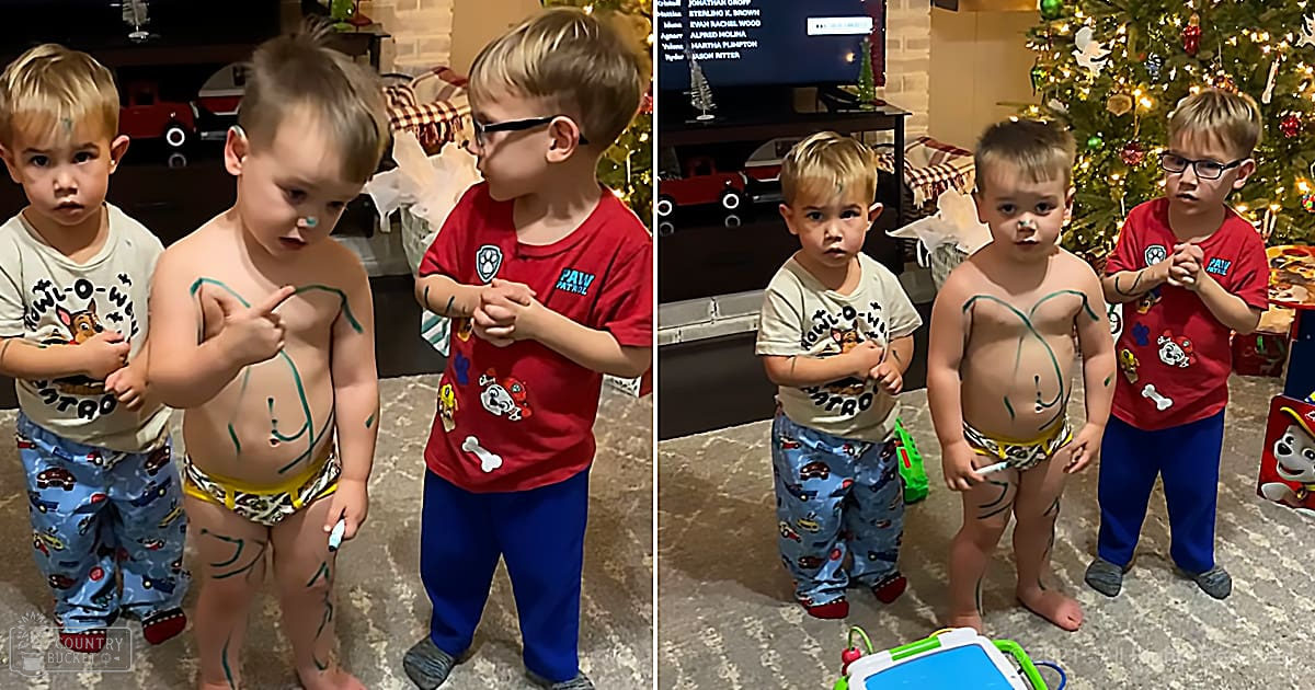 Three toddlers in casual home attire surrounded by Christmas decor, including a decorated tree and a guest list on television.