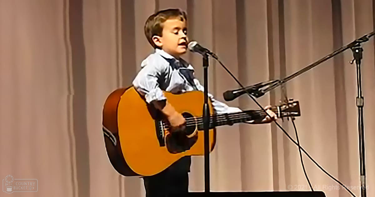 A young male child wearing a white shirt and dark pants performs music on stage, holding a guitar in front of microphones against light-colored curtains.