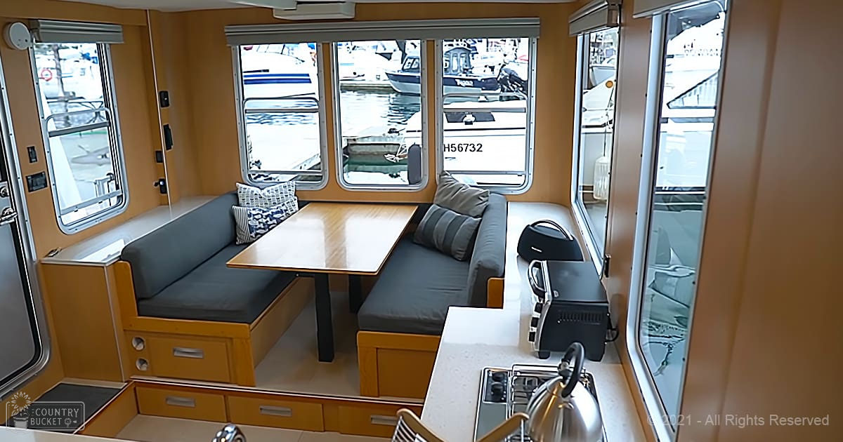 A compact boat cabin with built-in benches, cushions, a wooden table, storage drawers, and a countertop with a kettle, set in what appears to be a marina during the daytime.