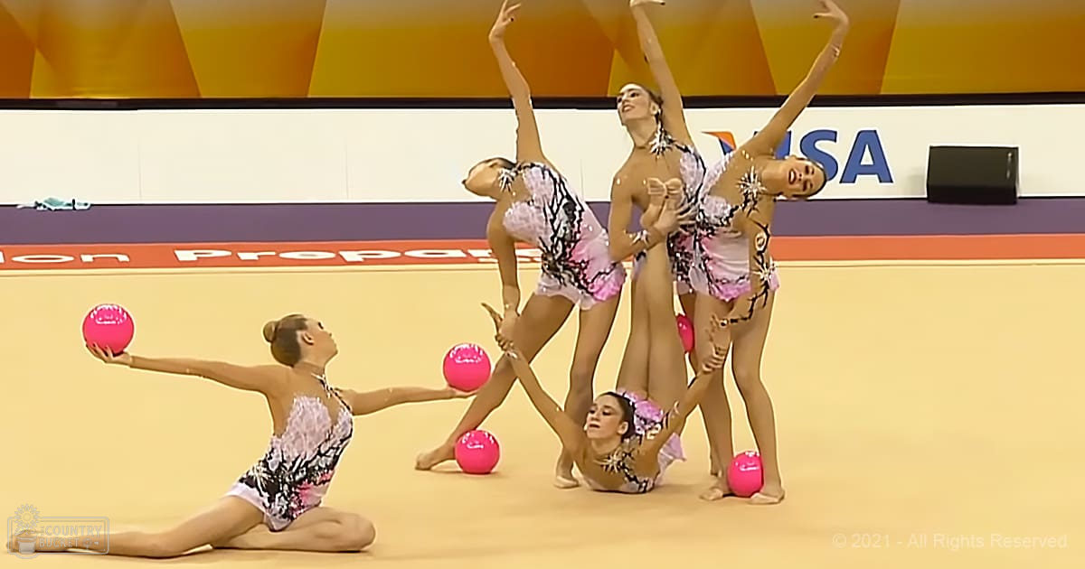 Young adult female rhythmic gymnasts performing with pink balls in a brightly lit indoor gymnasium, wearing patterned leotards and surrounded by logos on the backdrop.