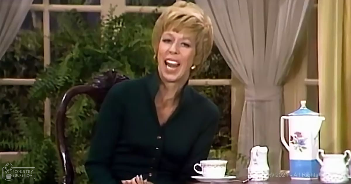 A middle-aged adult female in a relaxed, indoor setting, wearing a dark green cardigan with ornate furniture and home decor visible around her.