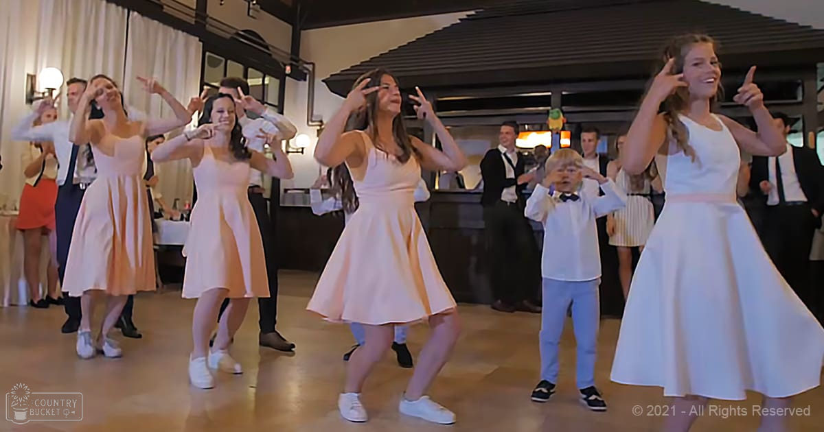 Group dance performance at an indoor event space with children wearing white dresses, adults in formal attire, and spring or summer evening lighting.