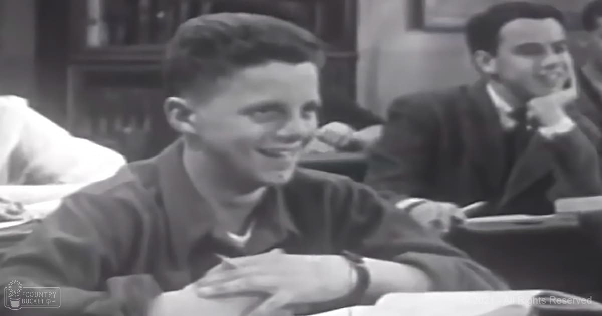 A young adult male child wearing a dark-colored shirt sits at a desk in an indoor classroom setting with another adult male seated nearby, suggesting a learning environment.