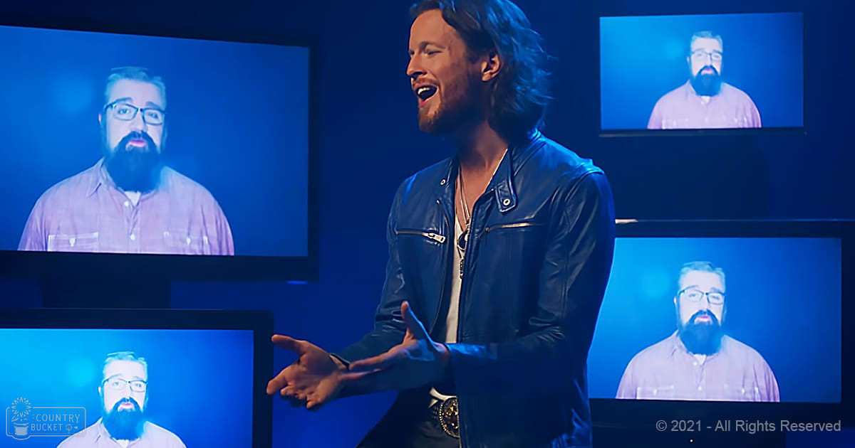 A young adult male stands indoors with screens displaying another bearded man. The setting is dark, illuminated by blue lighting.