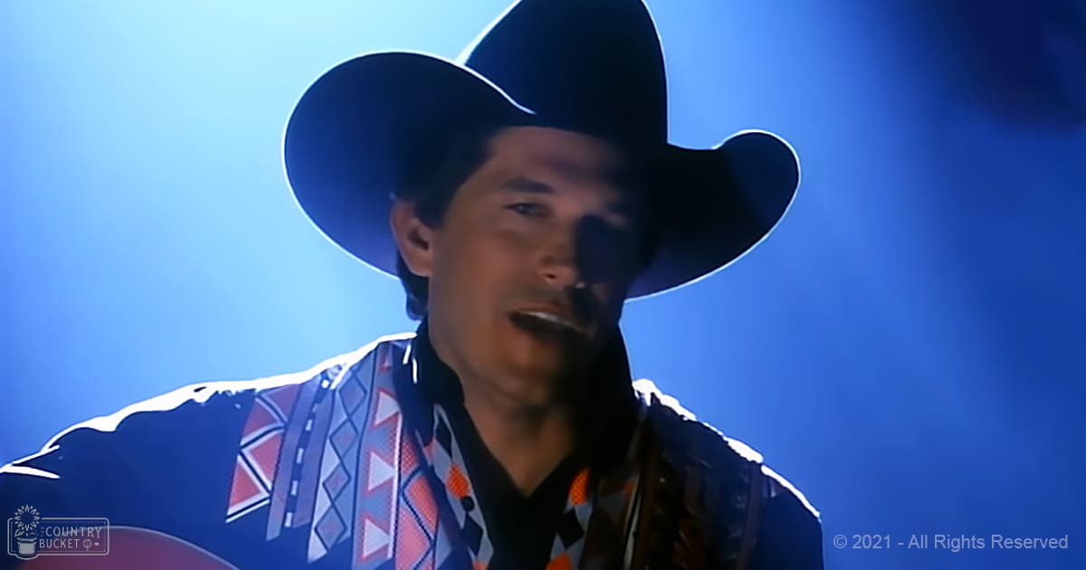 An adult male performer wearing a wide-brimmed black cowboy hat and a patterned shirt with geometric designs, partially visible on stage under blue lighting during an indoor concert.