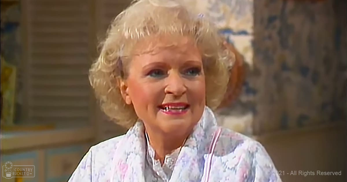 An older adult female wearing a loose, floral-patterned white blouse with long sleeves, sitting indoors near cabinets with glass doors; patterned curtain or wallpaper and beige or brown tones in the background suggest a home setting during daytime.