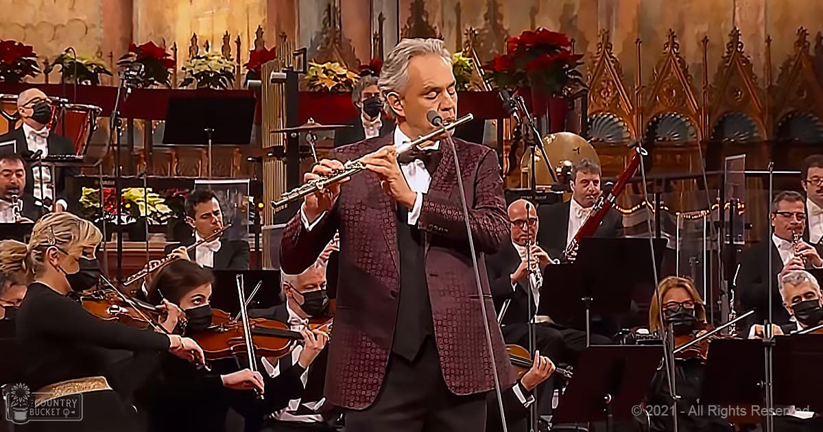 Adult male flutist in a maroon velvet jacket with white shirt and black trousers, surrounded by other musicians in an ornate concert hall setting during the holiday season.