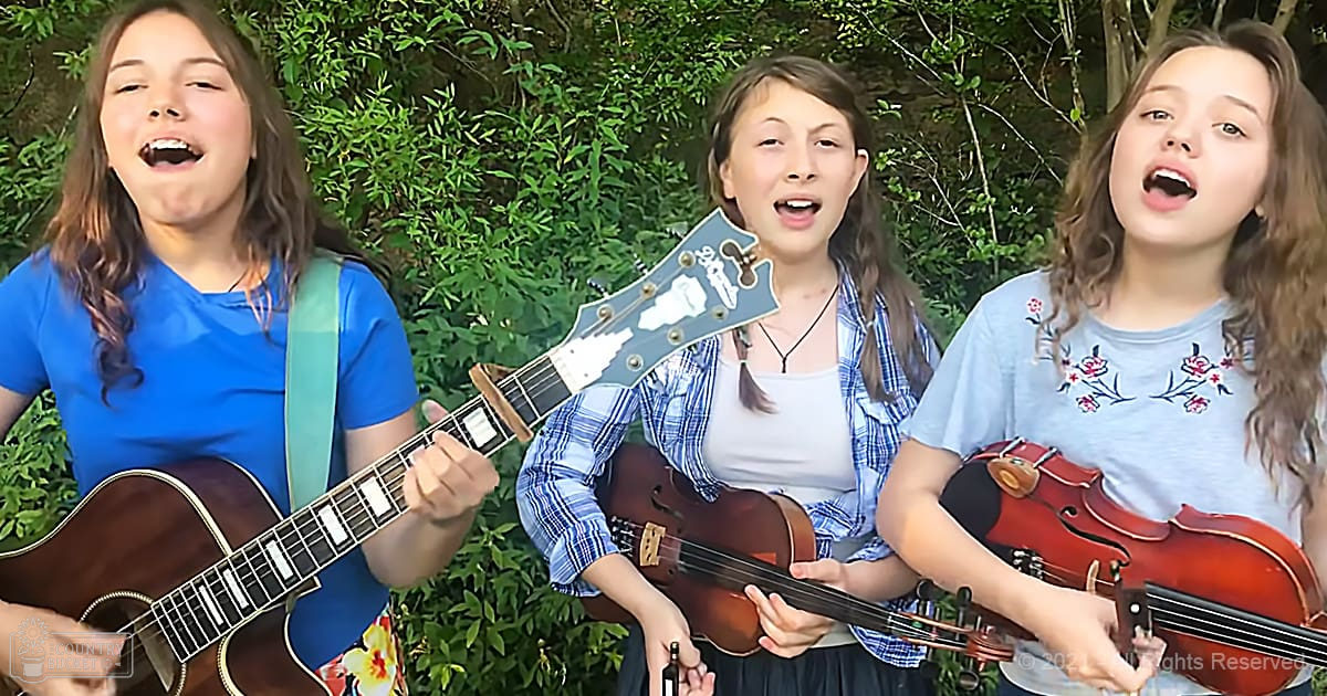 A group of young adults singing outdoors in a lush, green natural setting with musical instruments visible.